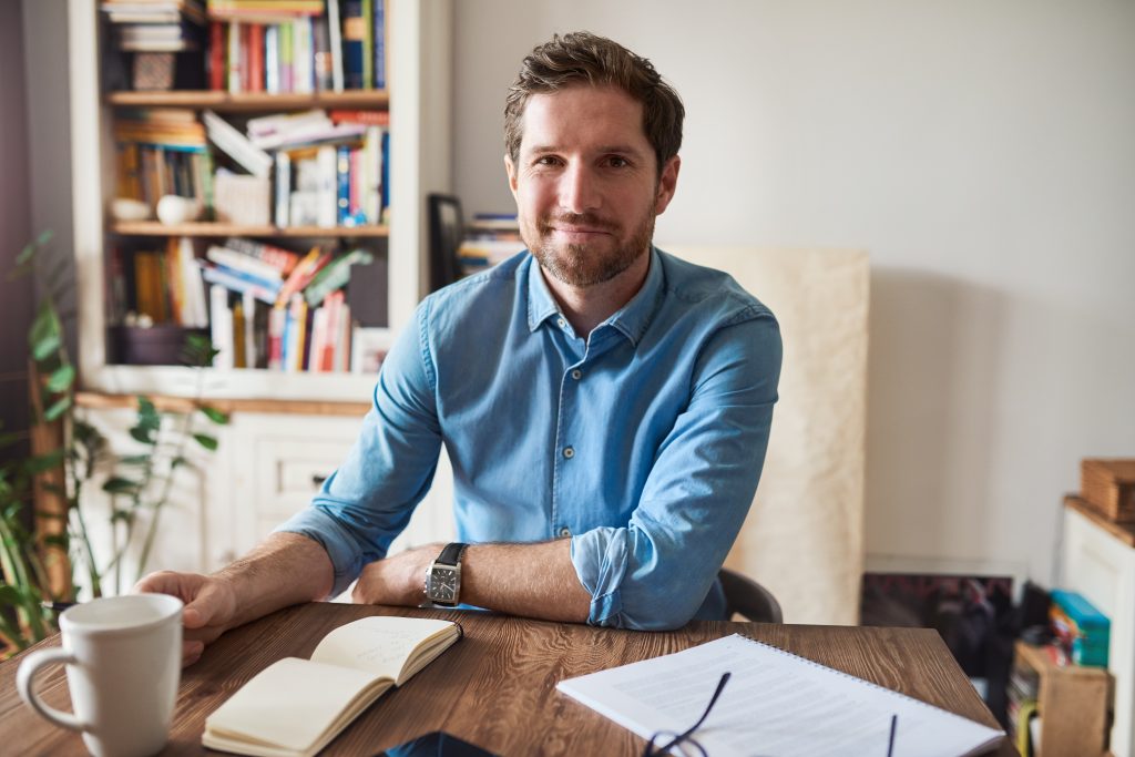 A man with short, dark hair and a beard is sitting at a wooden table. He is wearing a light blue button-up shirt and a wristwatch. On the table are an open notebook, a white coffee mug, and a pair of glasses. Behind him are bookshelves filled with books and various items, symbols of his self-published success. BookSelf Book Cover Design & Premade Book Covers