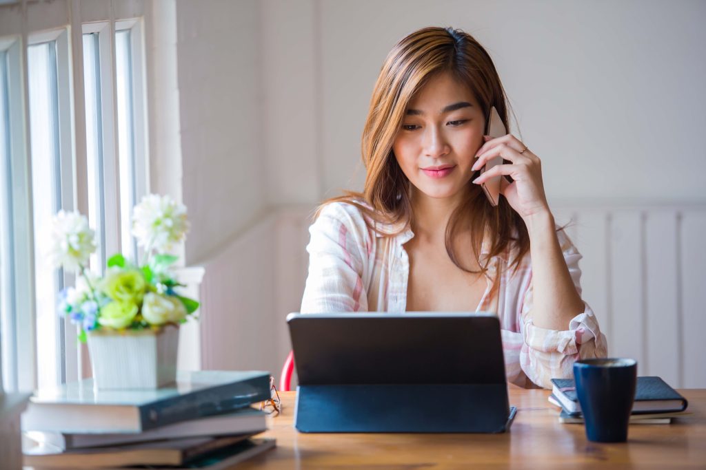 A woman with long hair sits at a wooden table in a bright room, engrossed in her tasks. She uses a tablet while holding a phone to her ear, possibly discussing self-publishing services for authors. On the table are a potted plant, a stack of books from BookSelf UK, a notebook, and a mug. Natural light streams through windows on the left, illuminating the scene. BookSelf Book Cover Design & Premade Book Covers